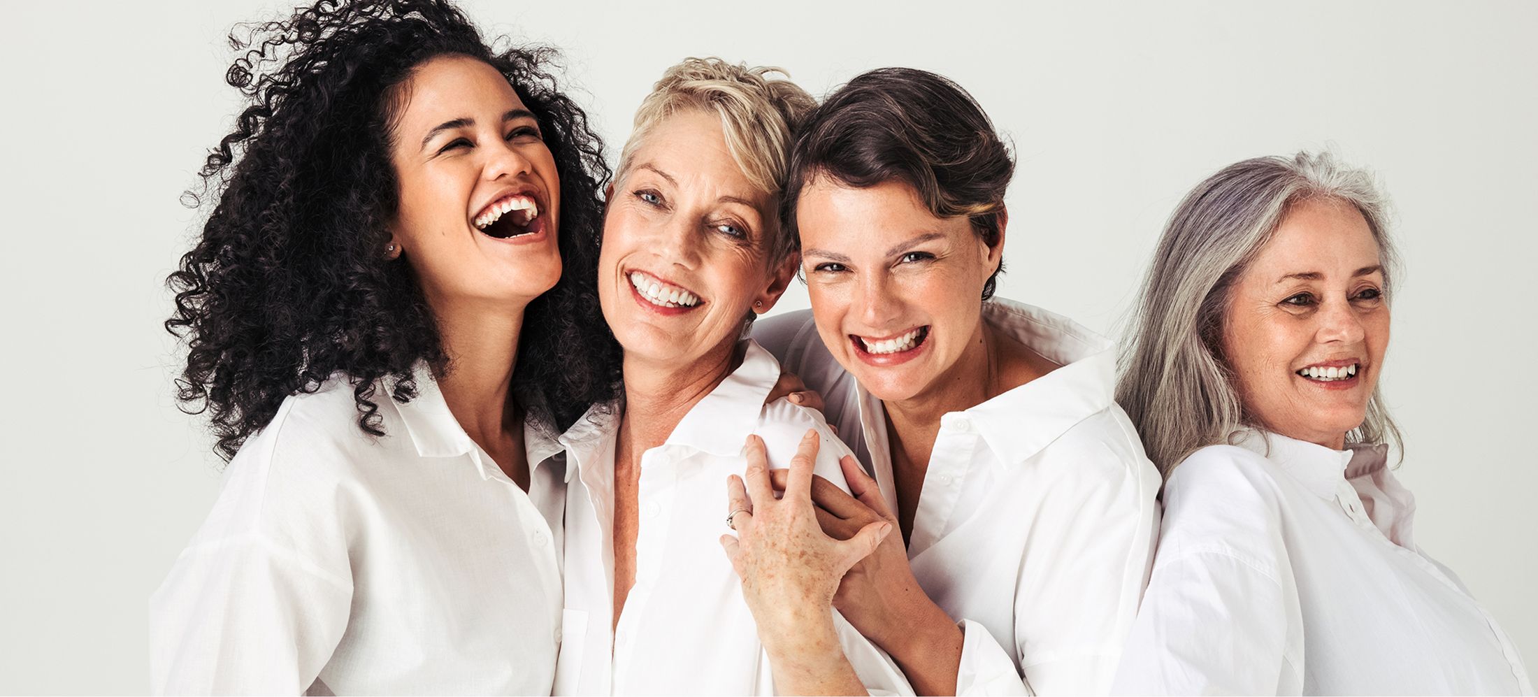 Four women laughing together in white shirts\